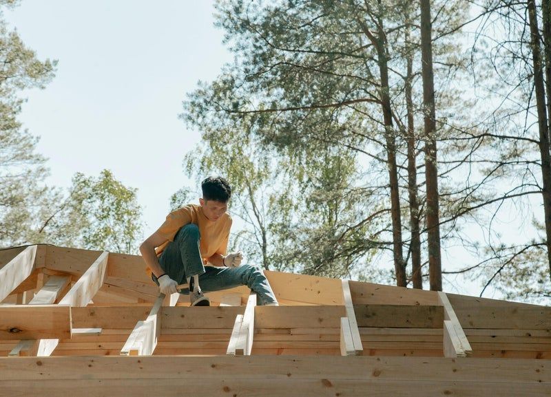 Ouvrier agenouillé sur une charpente de toit en bois sur un chantier de construction au milieu de grands arbres