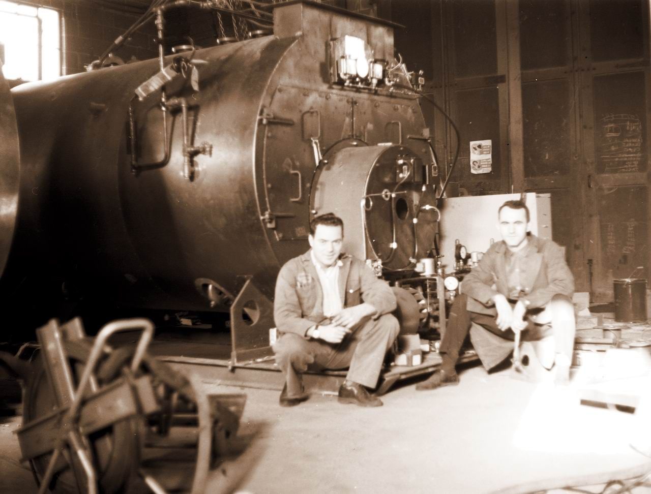 A black and white photo of two men sitting in front of a large machine