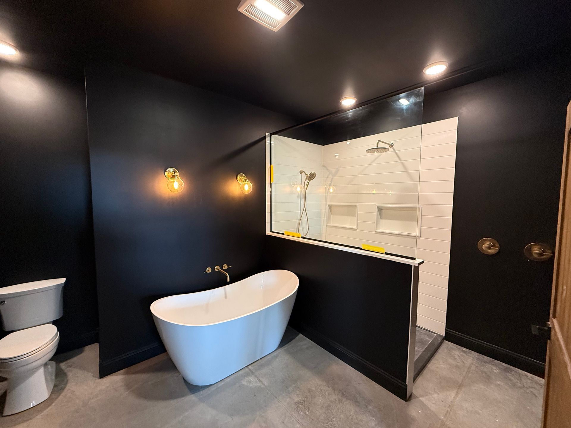 Black-walled bathroom with white bathtub, toilet, and shower with white tiled interior. Gold accents, overhead lighting.