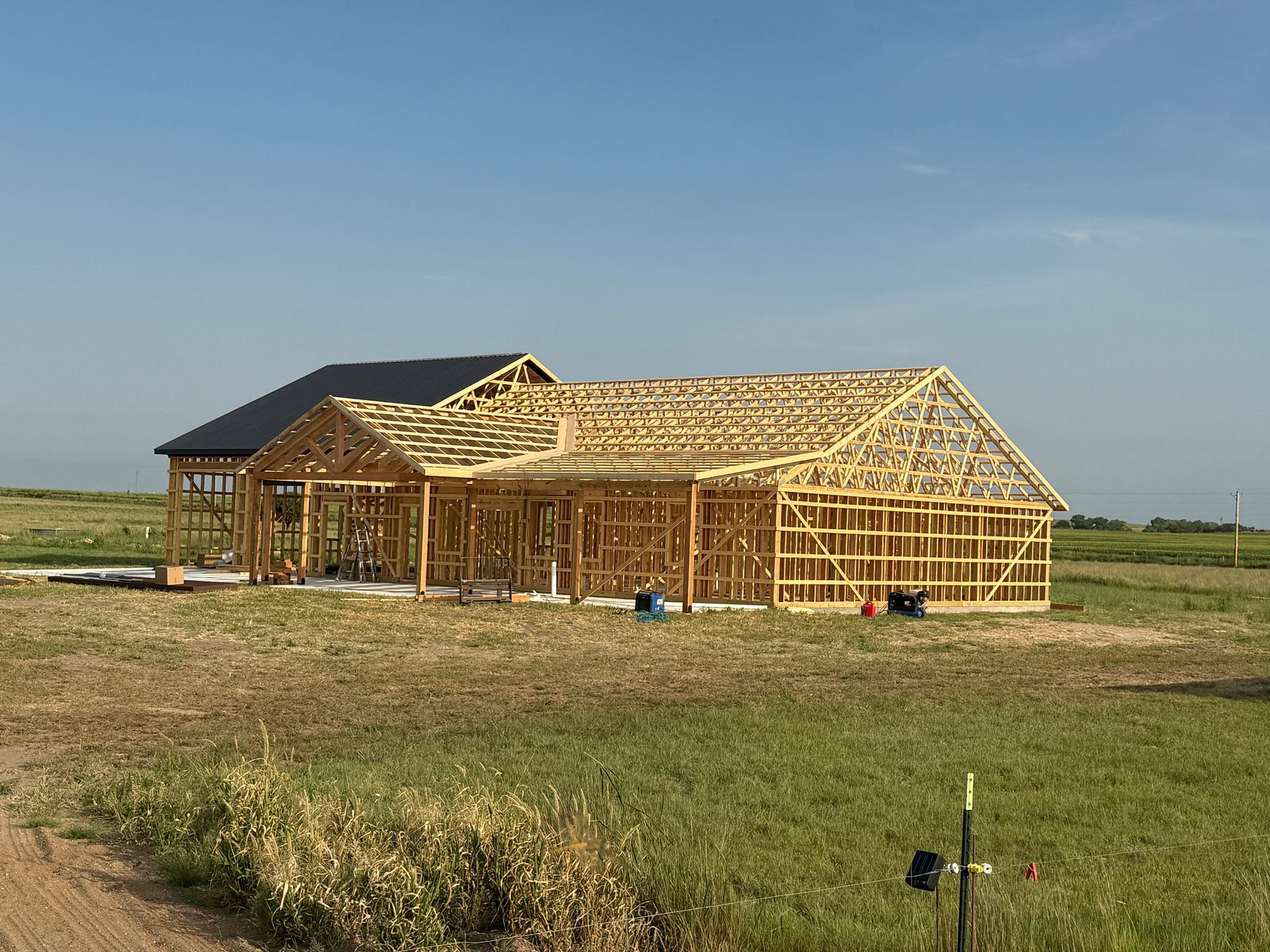 Wooden house frame under construction in a field, with a finished black roof visible in the background.