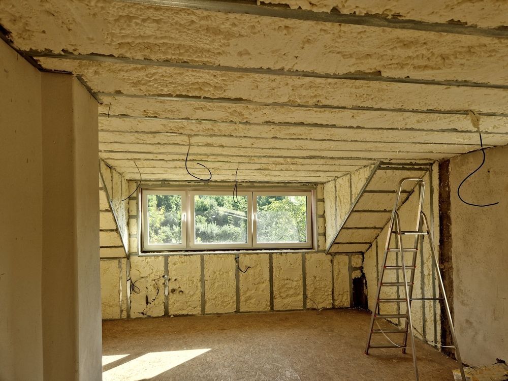 Interior room under construction, with insulated walls, exposed ceiling beams, a window, and a ladder.