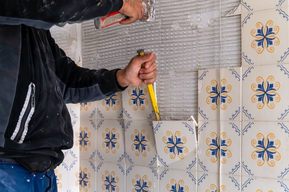 Person tiling a wall with blue and yellow patterned tiles, using a chisel and hammer.