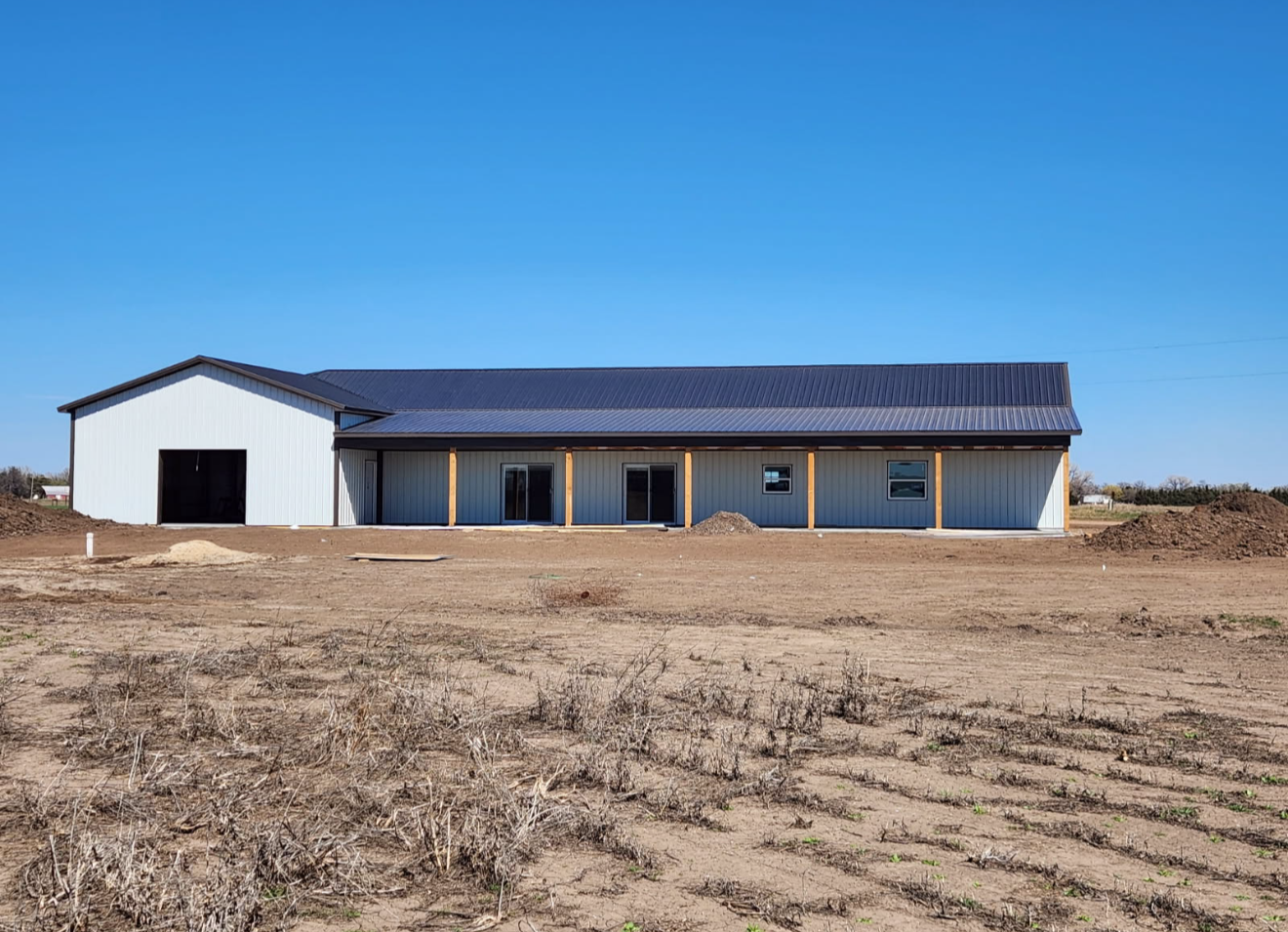 A light gray metal building with a dark roof and a covered porch on a dirt lot.