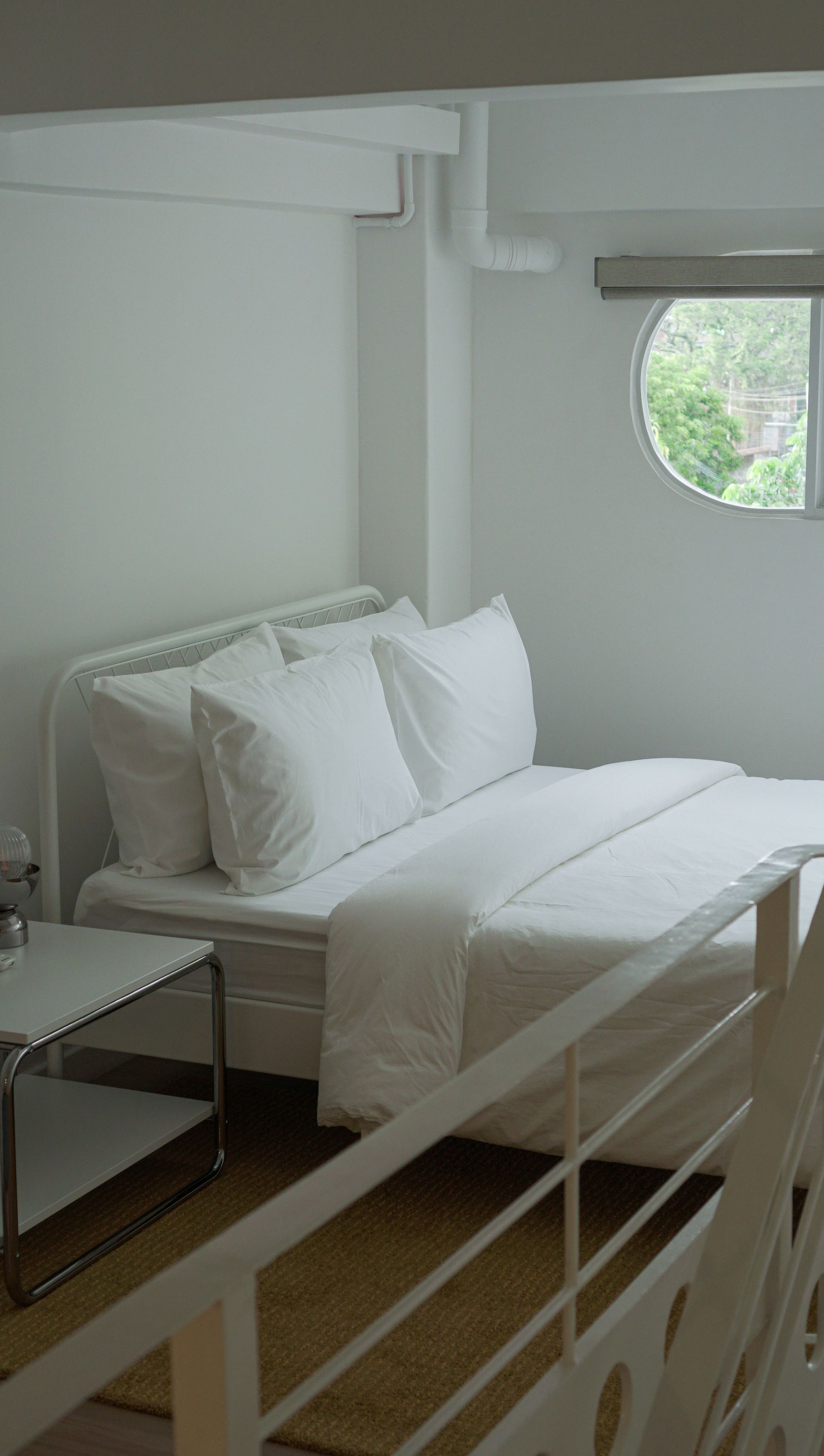 Bedroom with white bed, pillows, and nightstand; a circular window shows a tree-filled outdoor view.