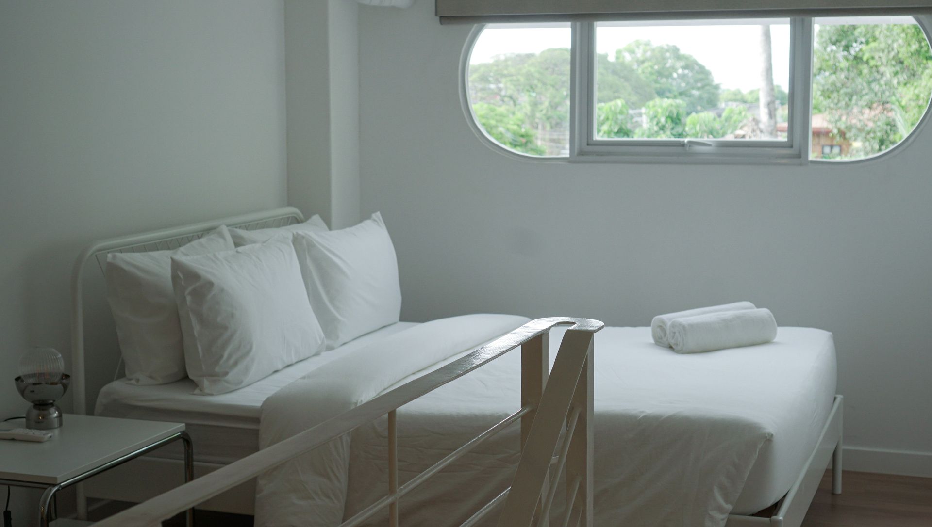 Bedroom with white bed, pillows, and a towel. A window reveals outdoor trees.