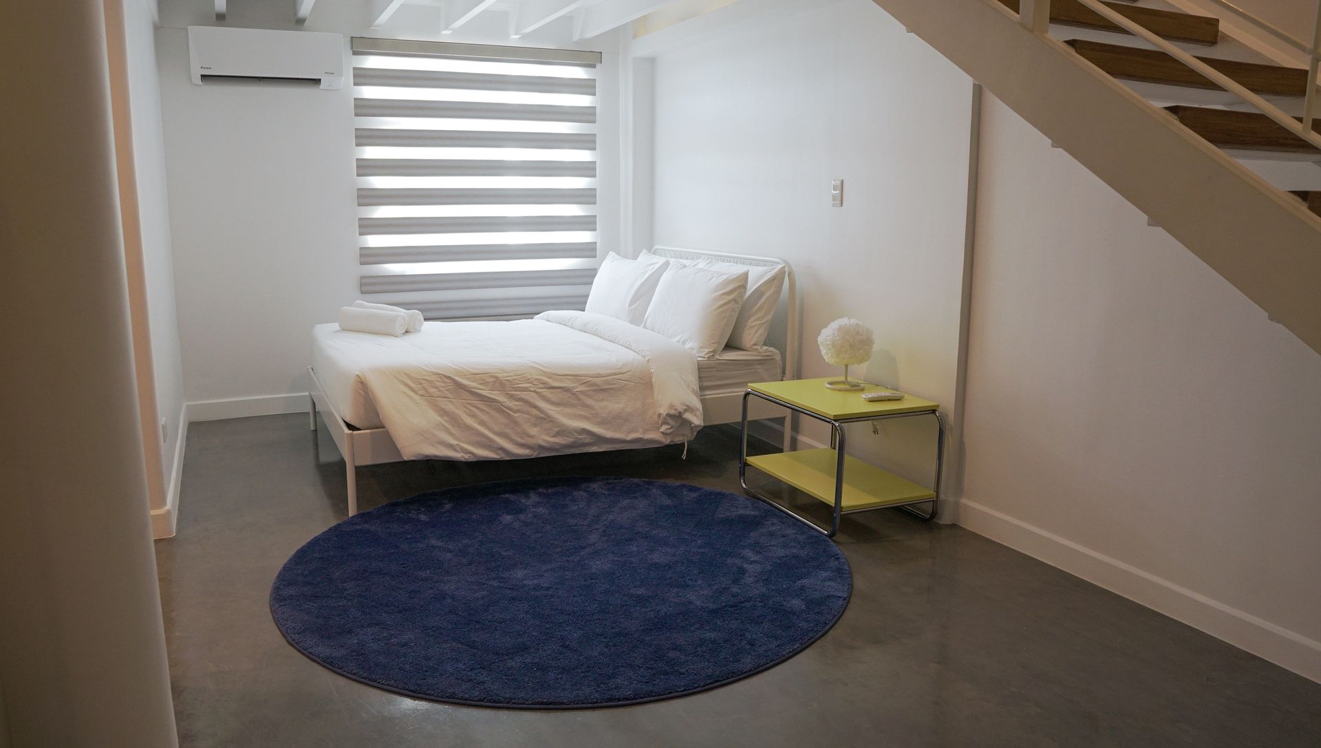 Bedroom with bed, blue rug, yellow side table, and window with blinds.
