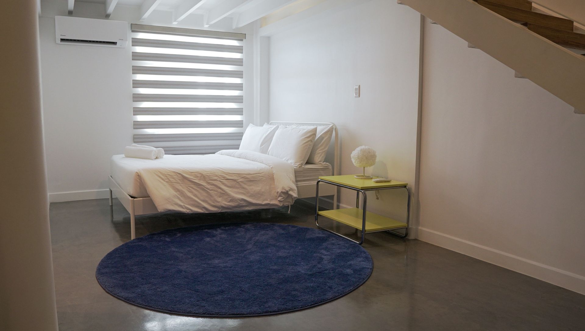 Bedroom with white bedding, blue rug, and small yellow side table. A window with blinds is present.