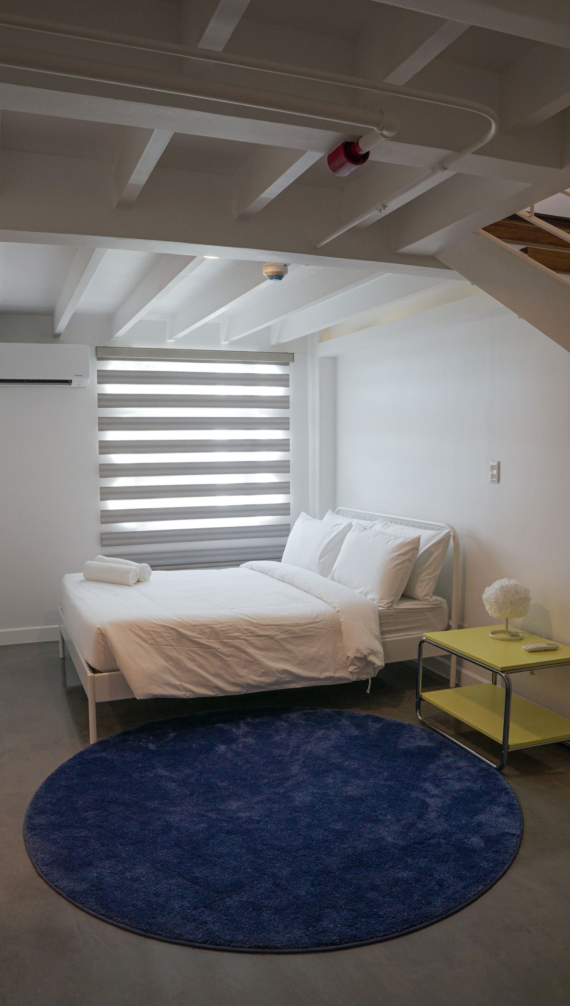 Bedroom with white bedding, blue rug, yellow side table, and window with blinds.