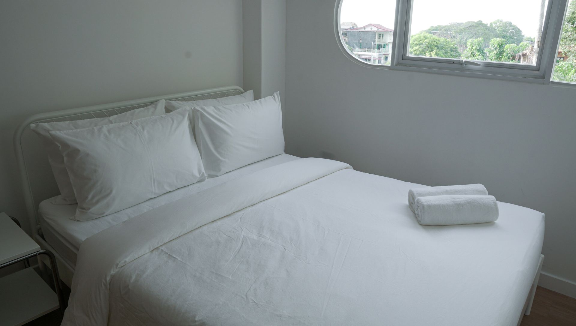 White bed in a bright room with a small window, two pillows, and rolled towels.