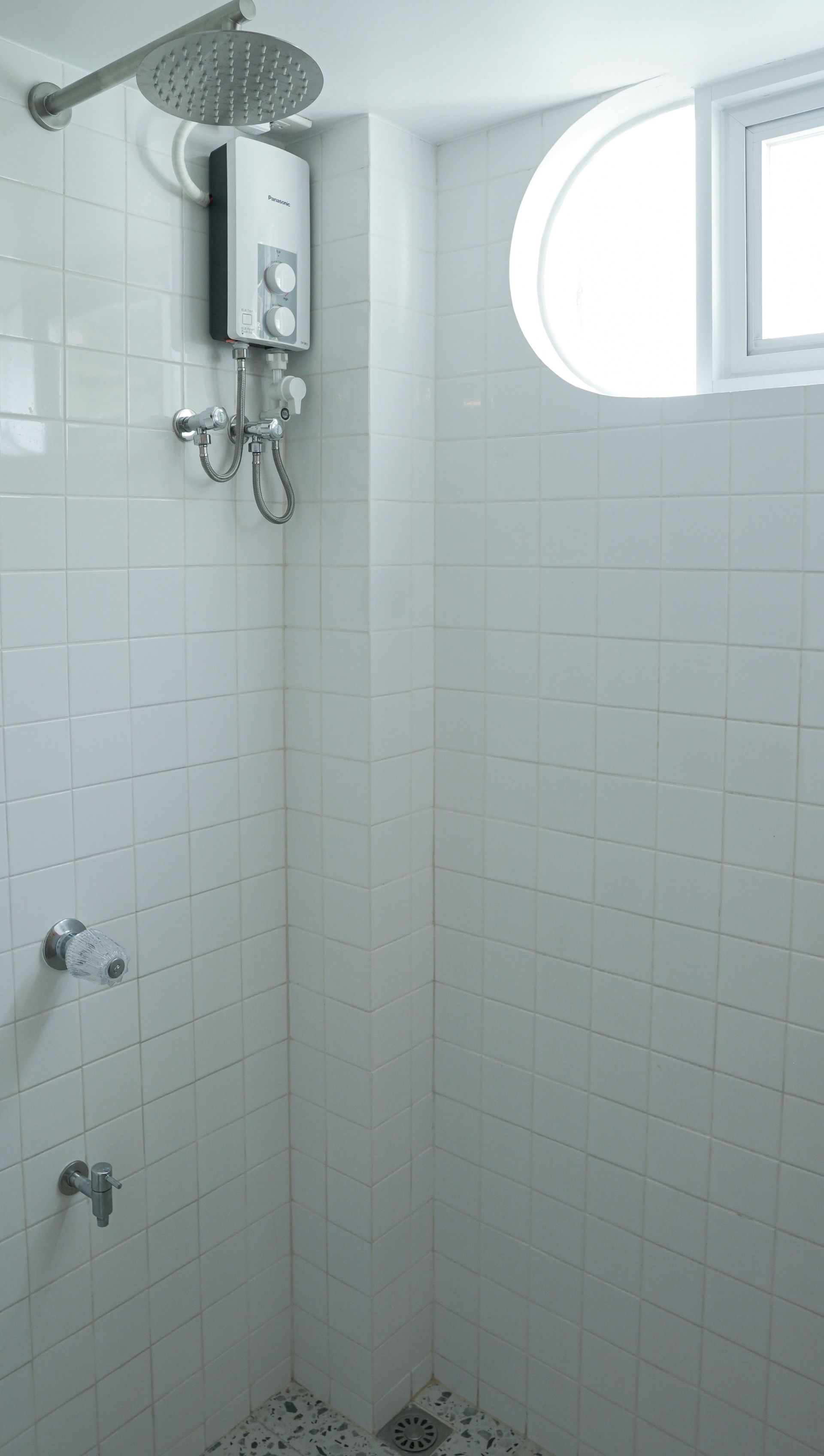 White tiled shower with rain showerhead, water heater, and small round window.