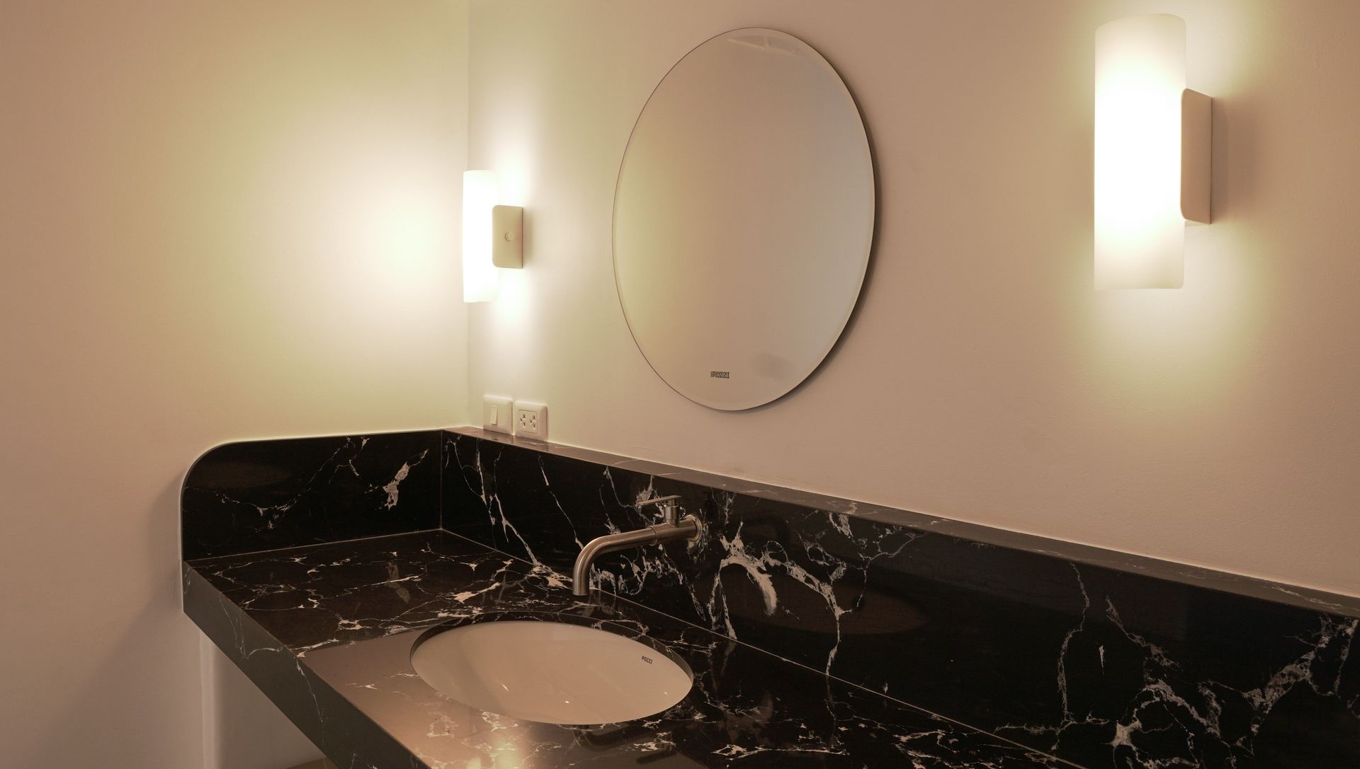 Black marble vanity with round sink, mirror, and two wall sconces.