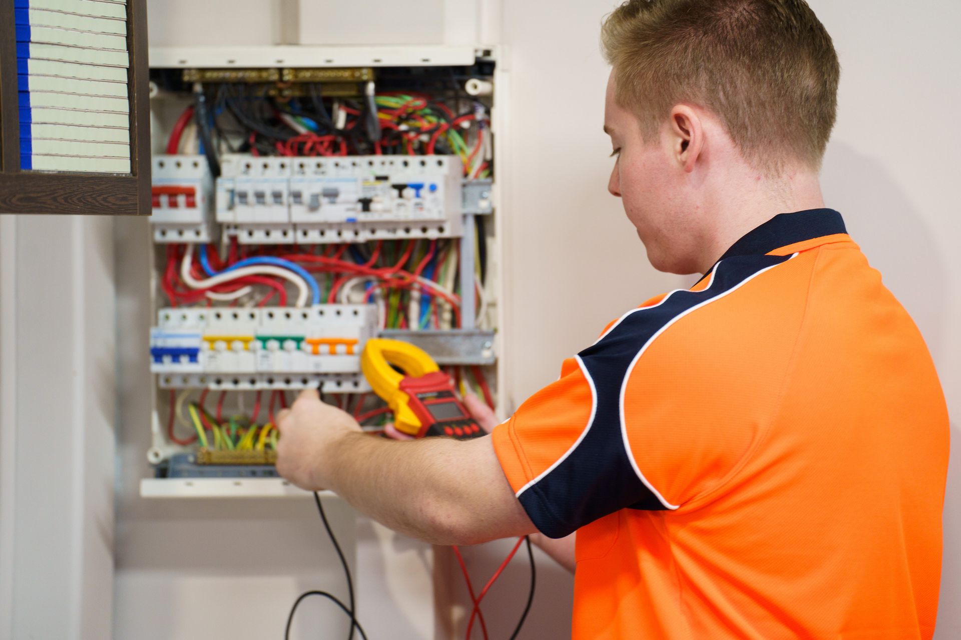 Electrician in orange shirt using a multimeter on an open circuit breaker panel.