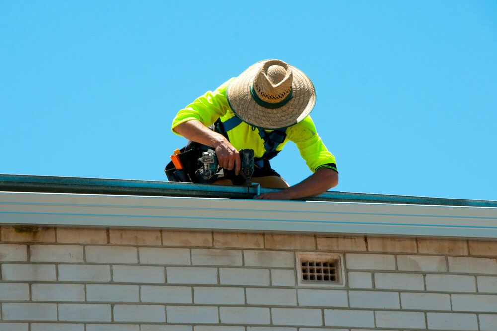 A Man Wearing a Straw Hat is Working on the Roof of a Building — Sheps Roofing Service Pty Ltd In Albion Park Rail, NSW