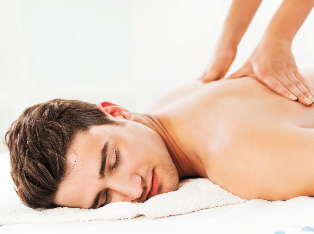 Ein Mann erhält in einem Spa eine Rückenmassage; er hat die Hände auf dem Rücken und liegt mit geschlossenen Augen auf einem Handtuch.