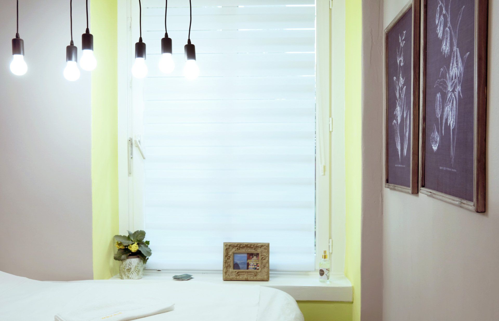 Schlafzimmer mit Fenster und Bett, Hängelampen, gerahmten Bildern und einer kleinen Topfpflanze.