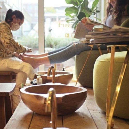 Eine Frau lässt sich im Spa die Füße pflegen, während eine andere entspannt eine Zeitschrift liest.