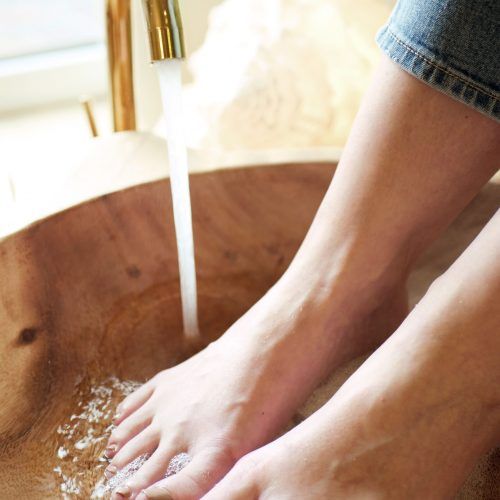 Füße in einer Holzschale, aus der Wasser aus einem goldenen Wasserhahn fließt; Person in Jeans.