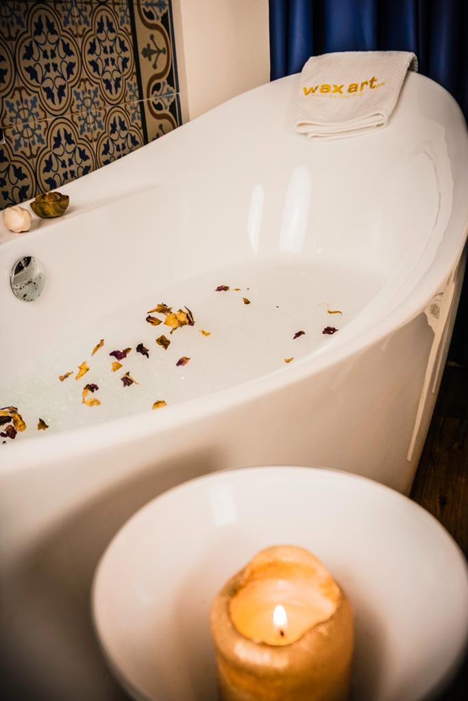 Weiße Badewanne mit Wasser und Blütenblättern, eine brennende Kerze und ein Handtuch.