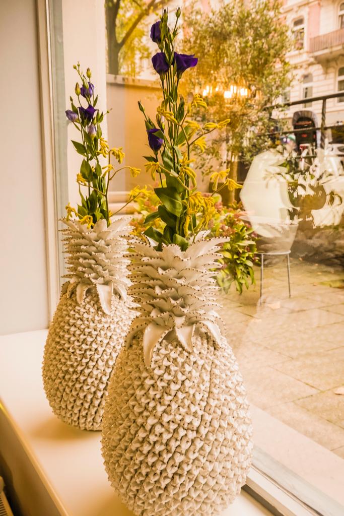 Auf einem Fensterbrett stehen zwei ananasförmige Vasen mit Blumen; durch das Fenster ist eine verschwommene Straßenszene zu sehen.