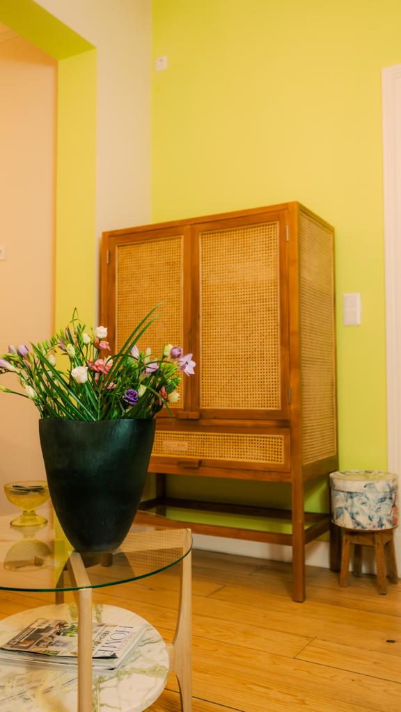 Holzschrank mit geflochtener Front, Blumenvase auf einem Glastisch und Hocker vor gelber Wand.