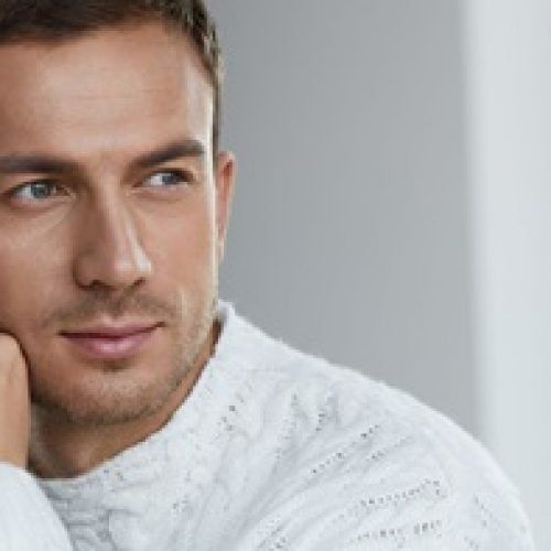 Ein Mann in einem weißen Pullover blickt nachdenklich nach links und stützt sein Kinn auf die Hand.