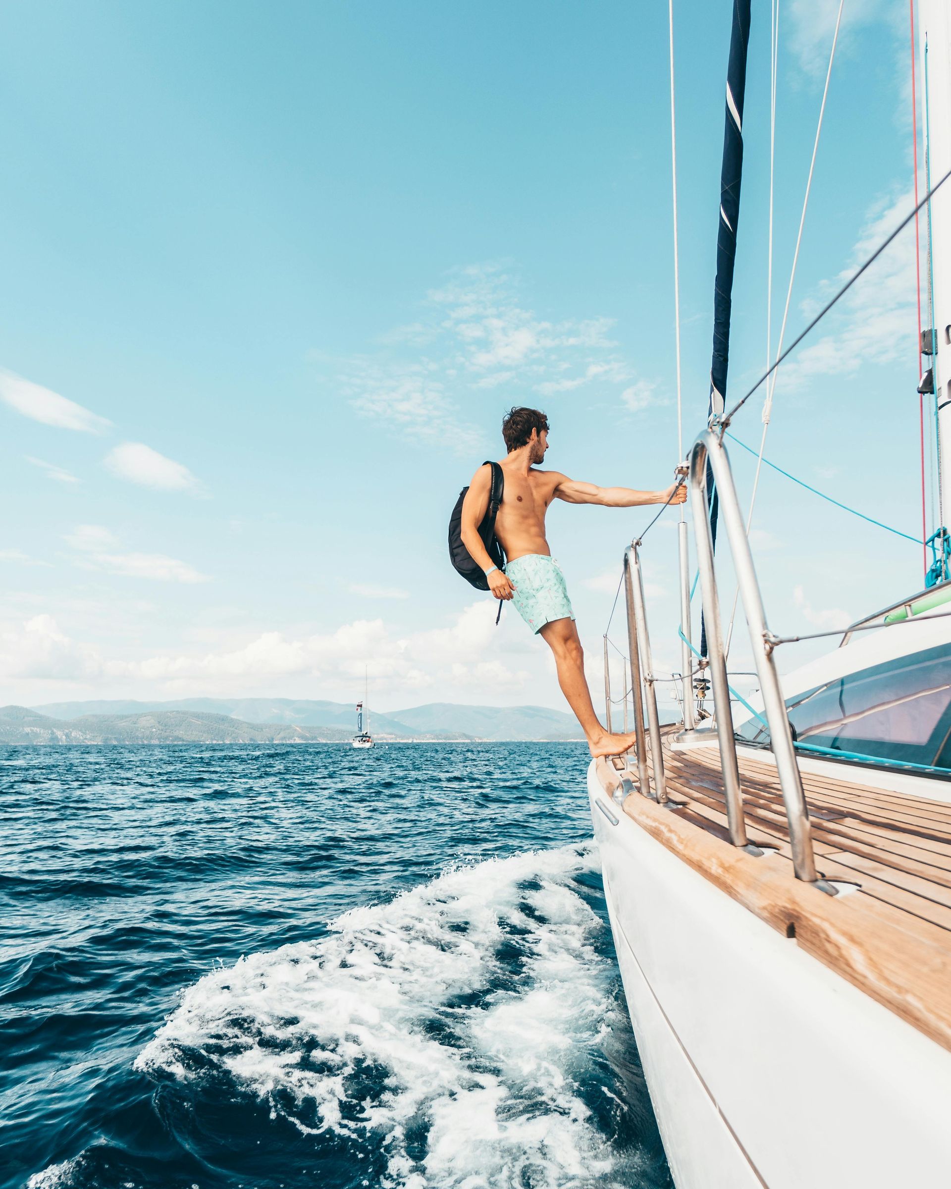 Mann auf einem Segelboot, an die Reling gelehnt, Rucksack auf dem Rücken, türkisfarbene Shorts, blaues Meer und blauer Himmel.