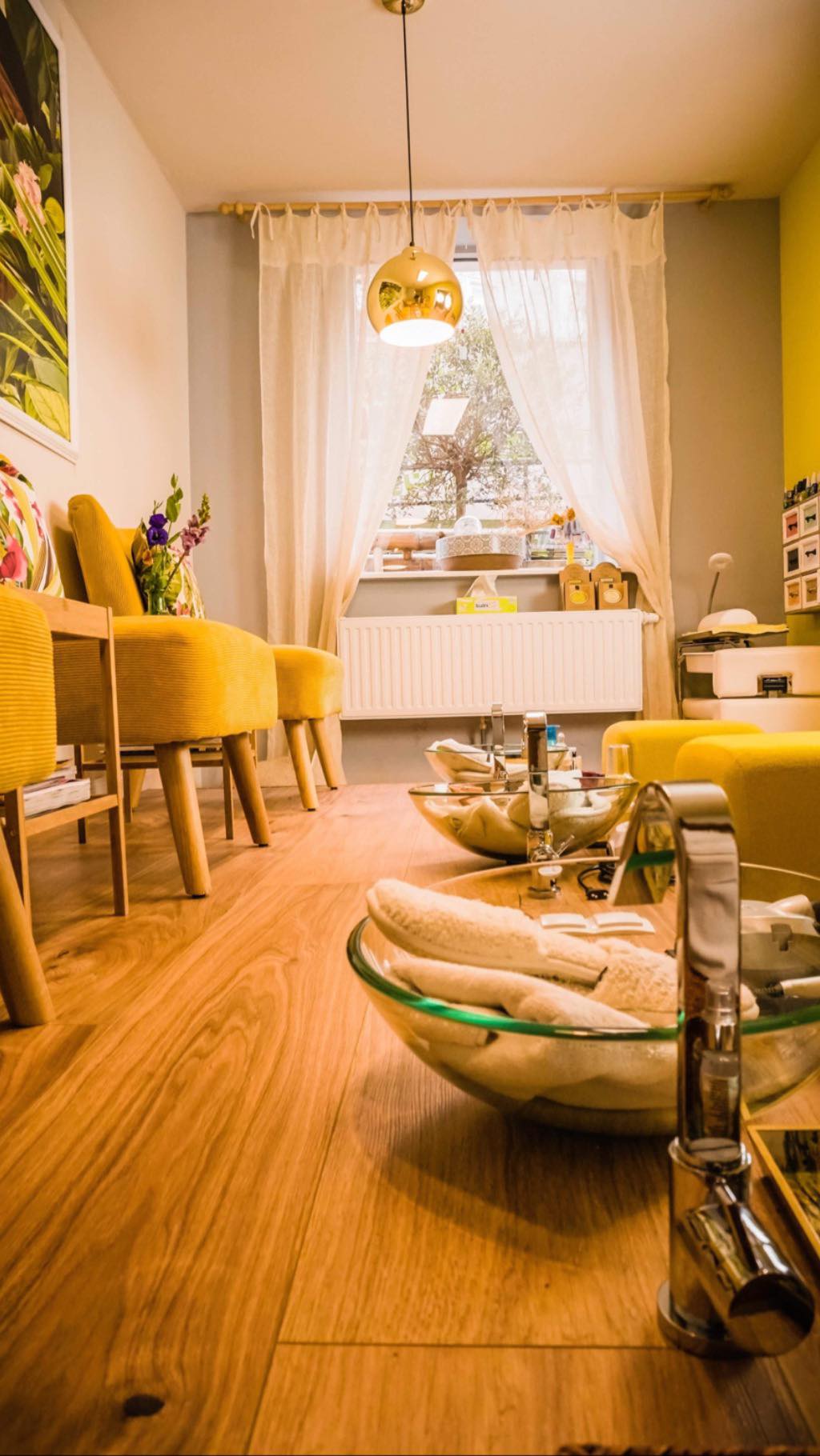 Gemütliches Nagelstudio mit gelben Akzenten: Stühle, Wände und eine Pendelleuchte. Schalen für Pediküren stehen auf dem Holzboden in Fensternähe.