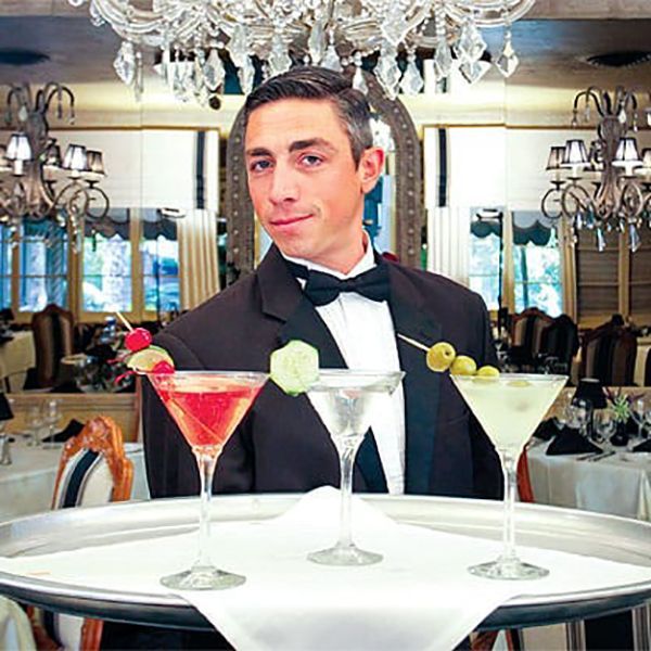Waiter in tuxedo holding a tray with three cocktails in a fancy restaurant.