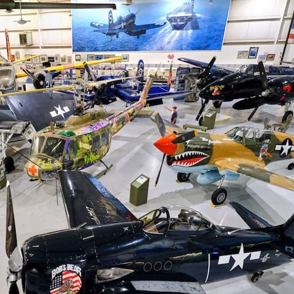 Inside a hangar, various vintage military aircraft on display. A man stands near a helicopter painted with camouflage.
