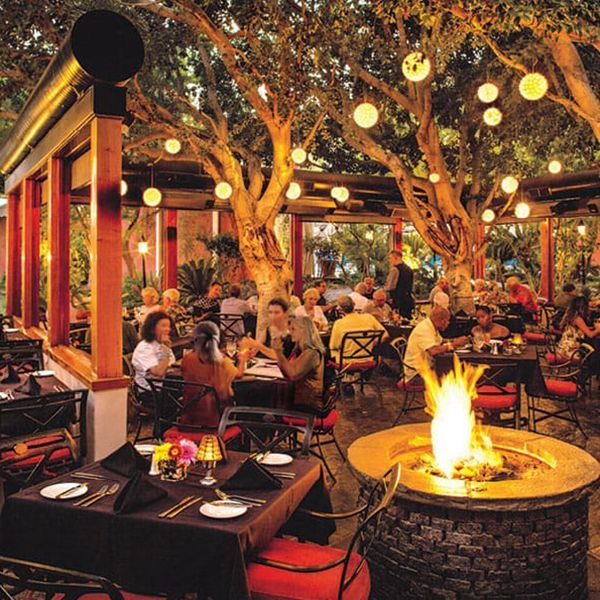 Outdoor restaurant seating under trees with glowing lanterns and a fire pit. People dine at tables.