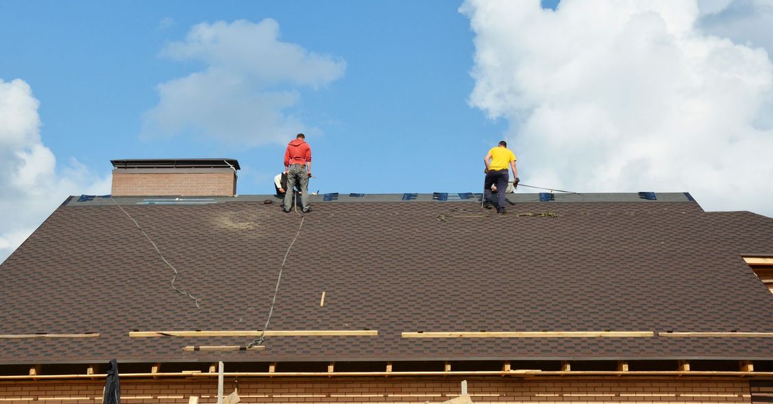 Two roofers in red and yellow jackets working on a shingled roof under a blue sky.