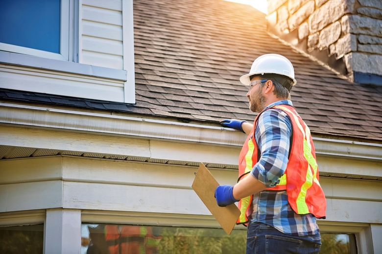 Inspector in hard hat and safety vest examining a house roof with a clipboard.
