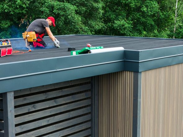Worker installing roofing on a flat roof beside a building with gray trim and green trees nearby