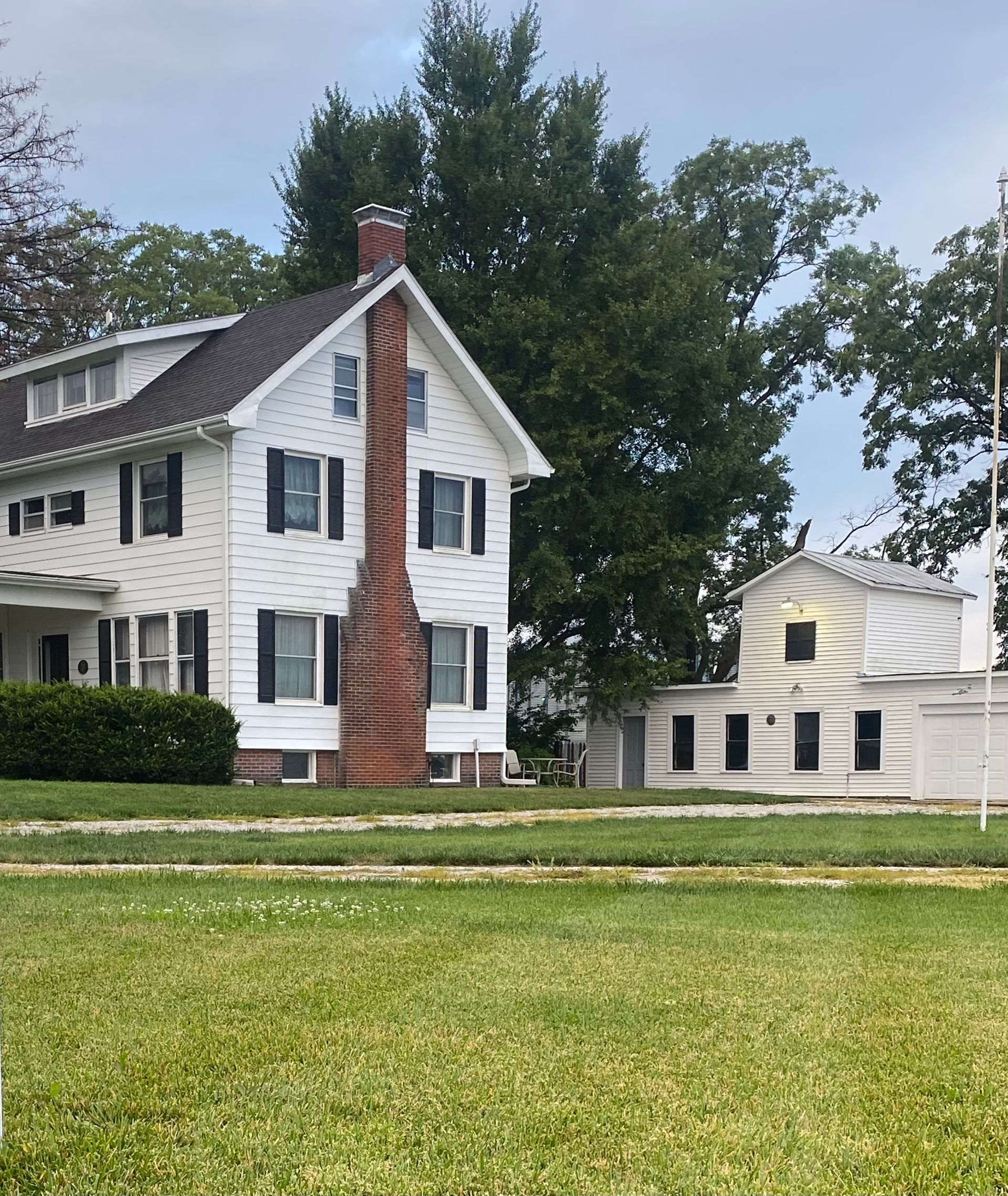 White farmhouse with black shutters and a brick chimney. Green lawn in the foreground, trees in the background.