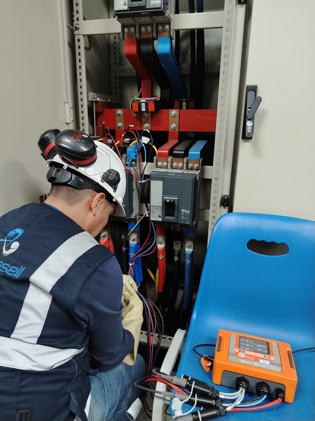Técnico inspeccionando equipos eléctricos en un armario, con cables azules y rojos y un medidor naranja cerca