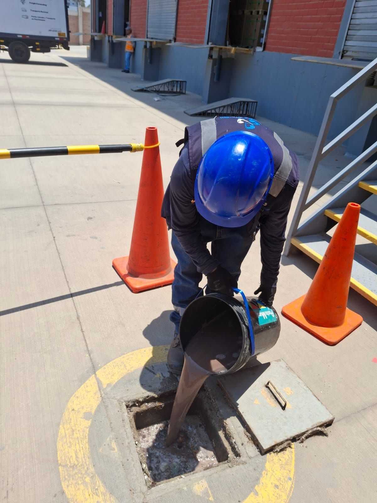 Un trabajador con casco azul inspecciona un desagüe de la acera junto a unos conos naranjas y una barrera de seguridad.
