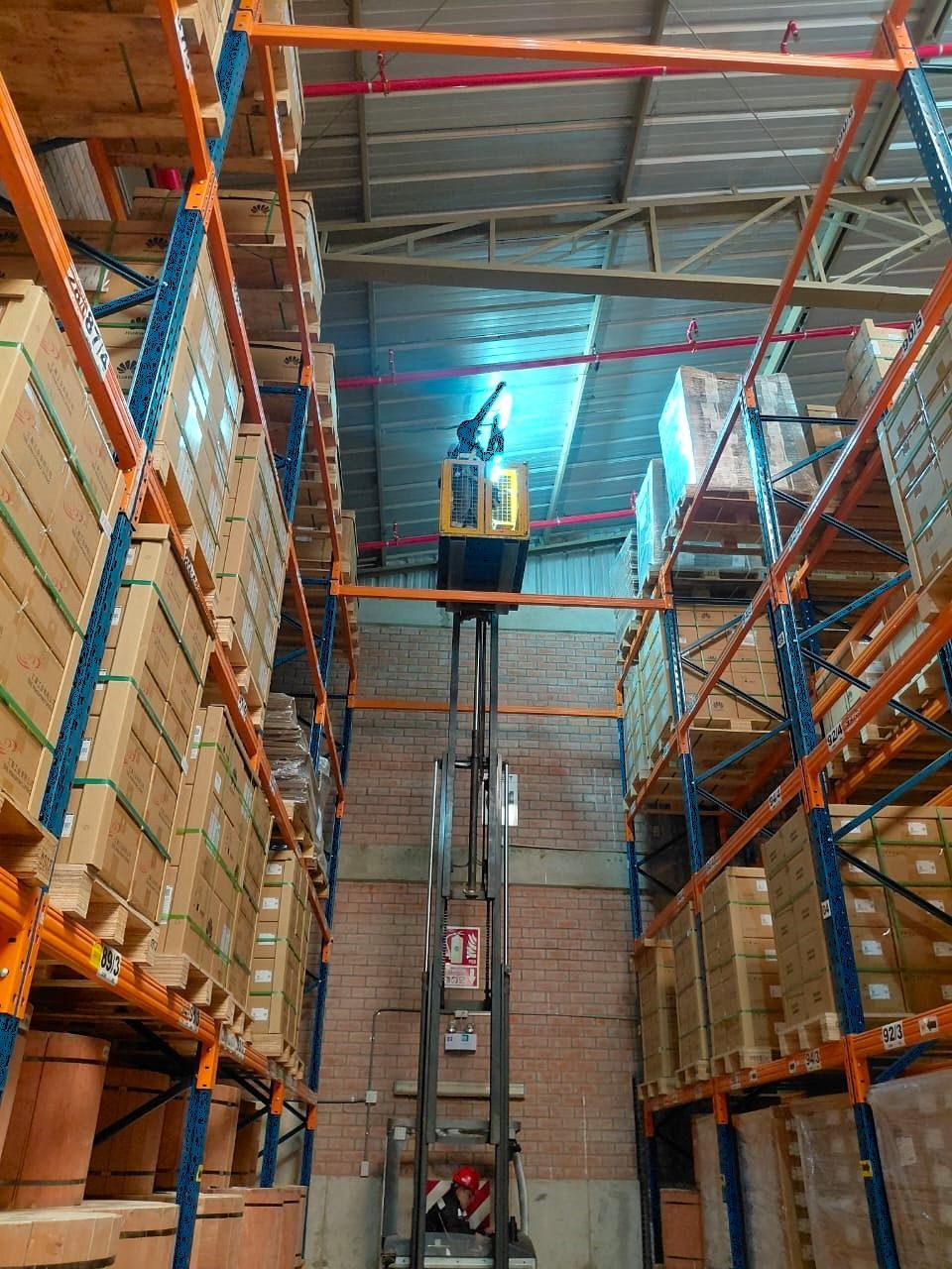 Un trabajador sobre una plataforma elevada en un pasillo de almacén, entre estanterías altas, iluminado por una luz azul intensa.