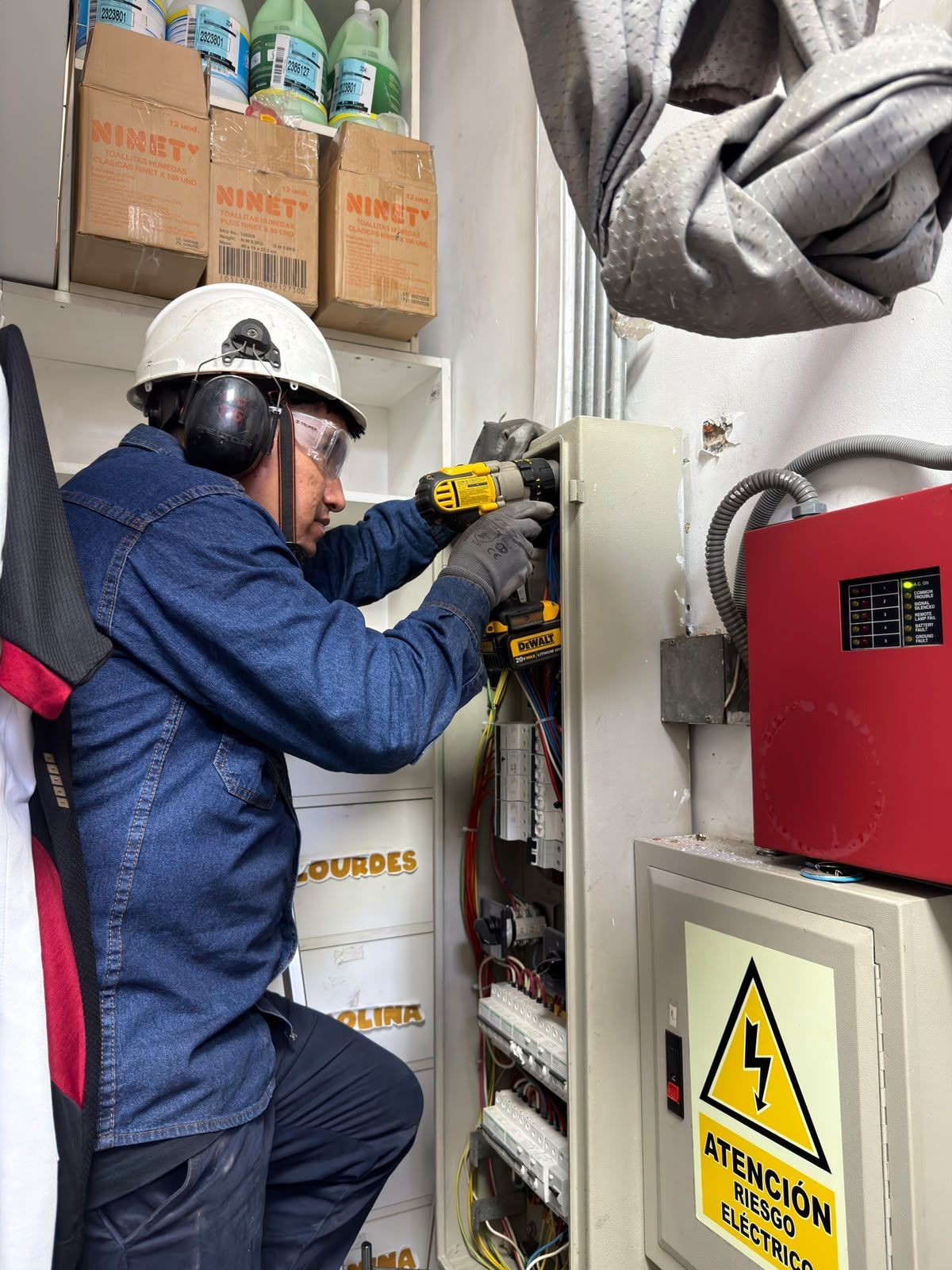 Un trabajador con mono azul ajusta un cuadro eléctrico junto a un equipo rojo y una señal de advertencia en una sala de servicios
