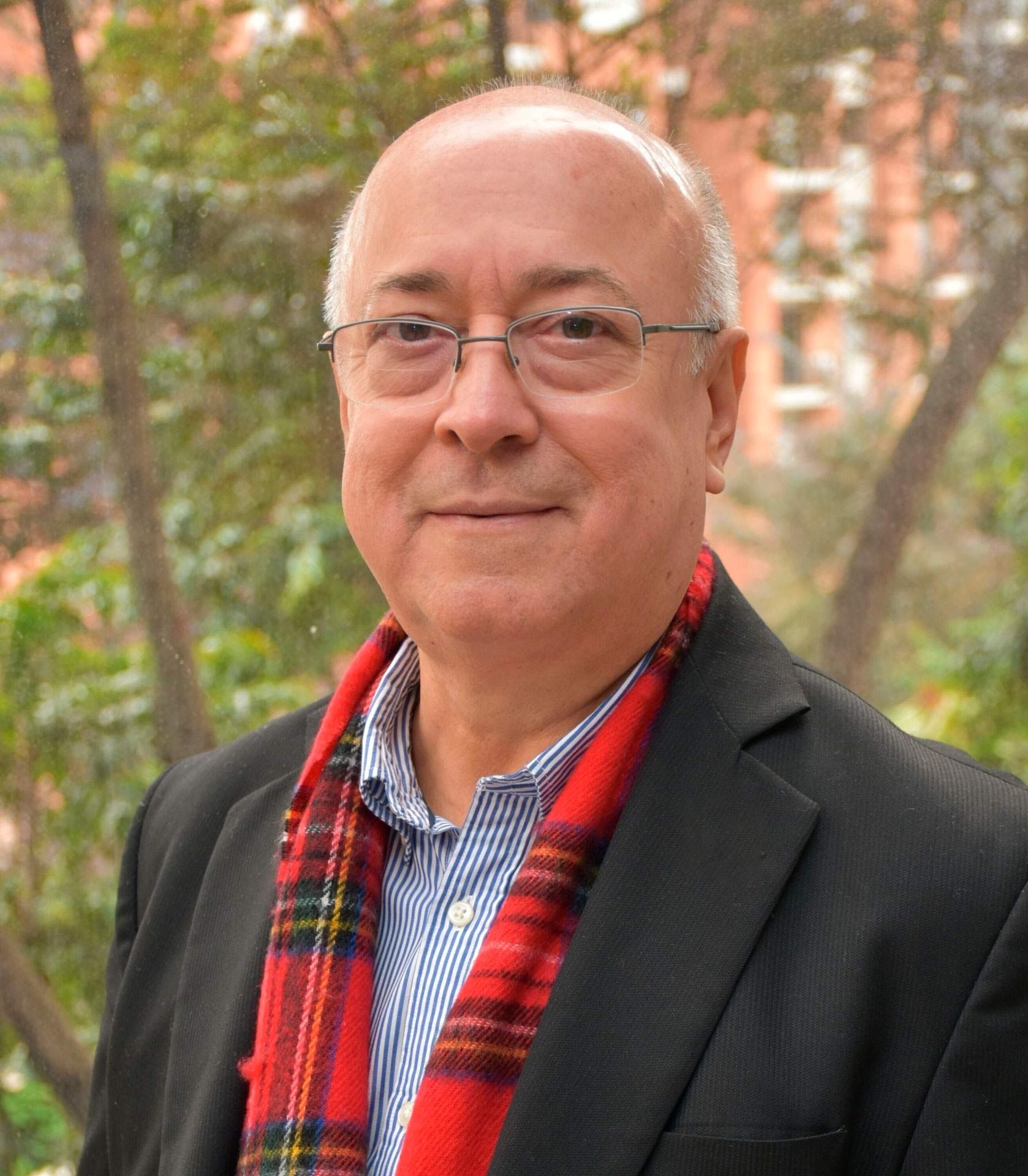 Una persona con gafas, una camisa a rayas, un blazer negro y una bufanda a cuadros roja, de pie al aire libre con árboles.