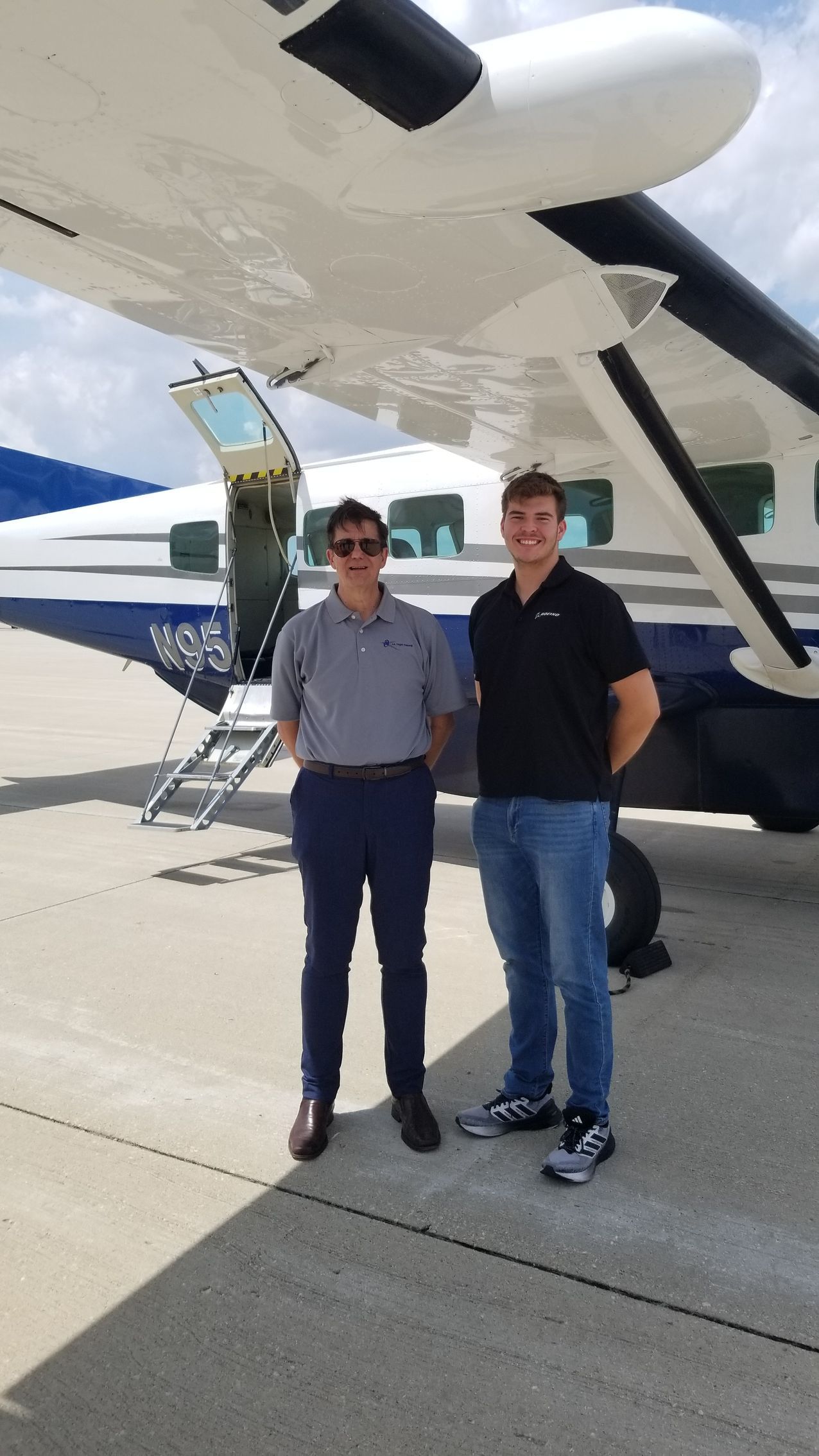 Two people standing beside a small airplane on a tarmac, under the wing, with the plane door open.