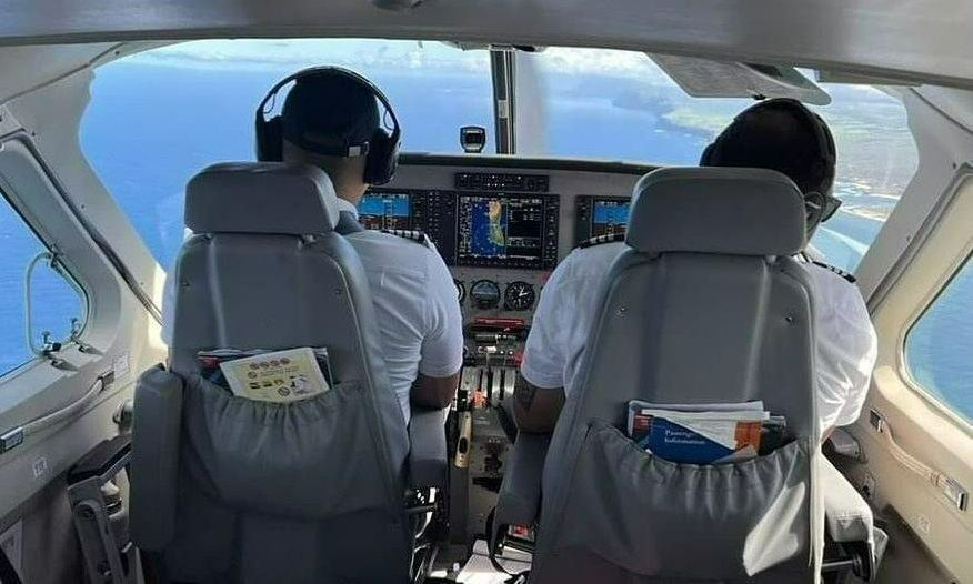 Two pilots flying a twin-engine aircraft with glass cockpit displays over the coastline.