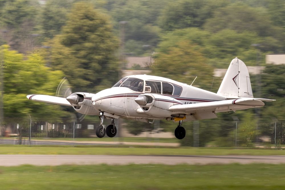 Small twin-engine propeller plane landing on a runway with landing gear down, trees blurred in the background