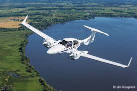 Diamond DA42 twin-engine training aircraft flying over lake and farmland landscape.