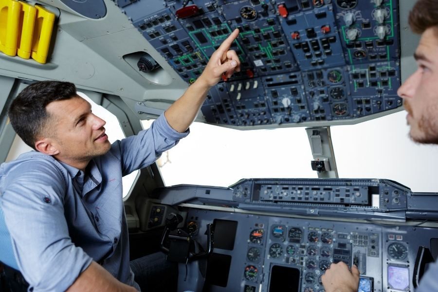 Flight instructor demonstrating overhead cockpit controls to a student in an airline simulator.
