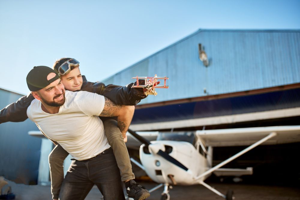 A father and child play pilot together outside a hangar.