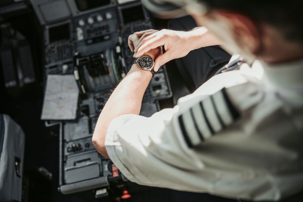 Pilot in cockpit checking wristwatch; instrument panel visible.