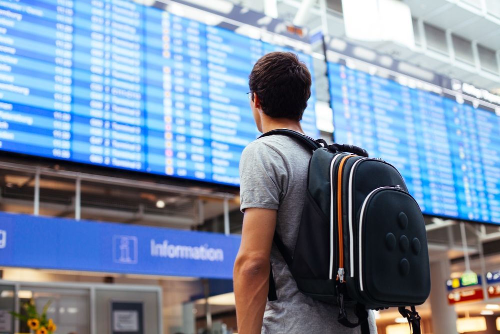 A man with a backpack is standing in front of an airport departures board with delayed flights due to rain.