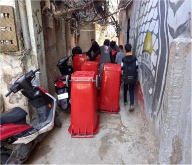 A group of people are walking down a narrow alleyway carrying red trash cans