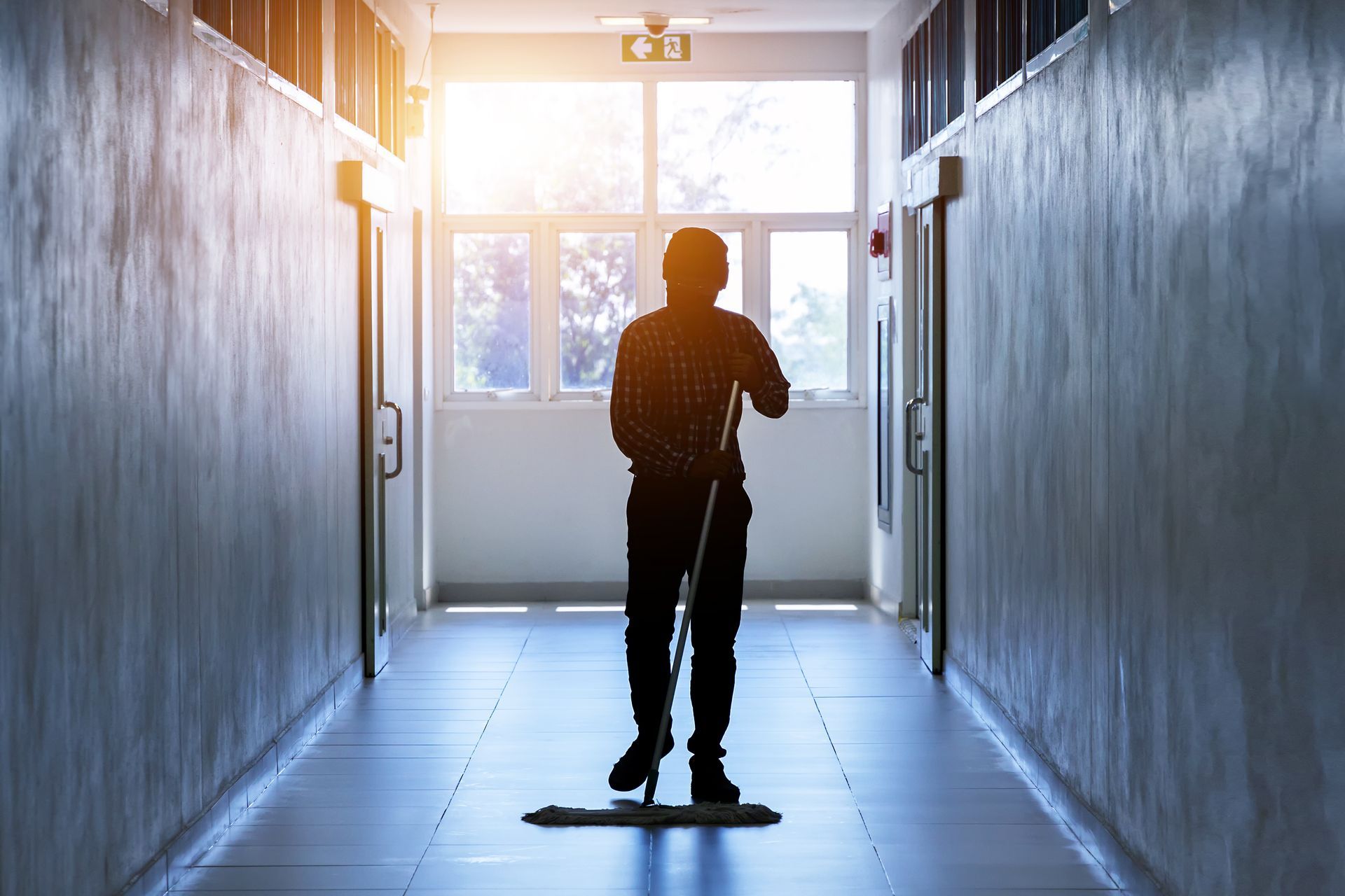 Person mopping a hallway with sunlight streaming through a window at the end.