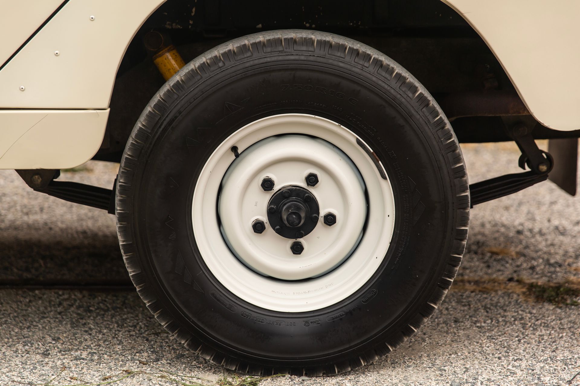 Close-up of a cream-colored vehicle’s rear wheel and tire on a gravel surface.